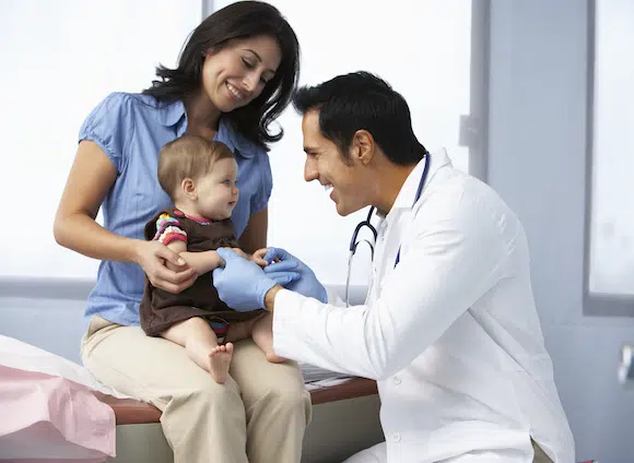 Doctor In Surgery Examining Baby Girl With Mother Smiling