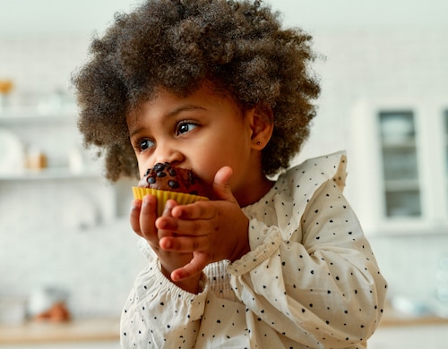 An African American little baby with curly fluffy hair in a dress eats a chocolate muffin