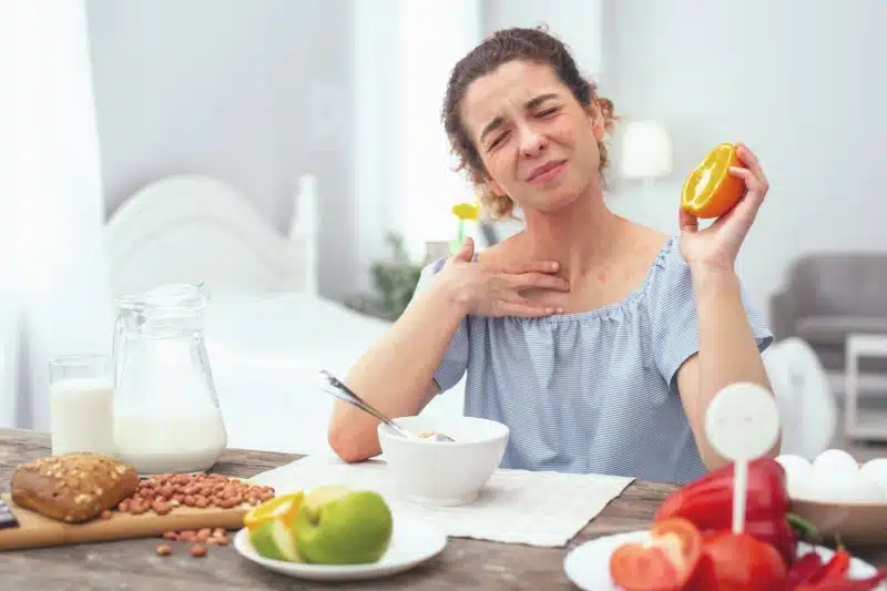Woman experiencing throat itchiness, a symptom of Oral Allergy Syndrome, while having yogurt and orange for breakfast at her bedroom.