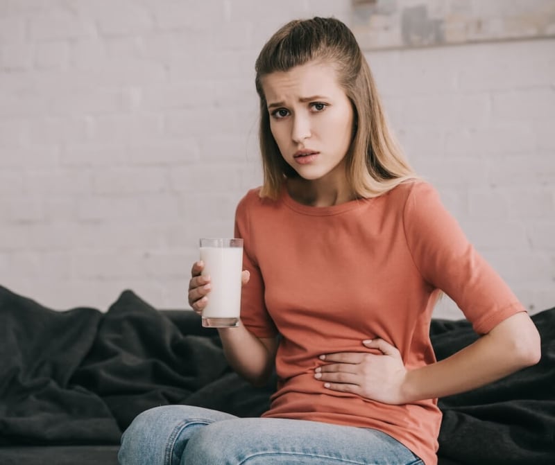 Upset blonde woman with lactose intolerance holding glass of milk while sitting on sofa grabbing her belly