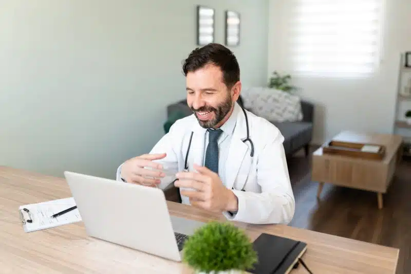 Friendly Hispanic doctor working from home and talking to a patient in a telemedicine consultation