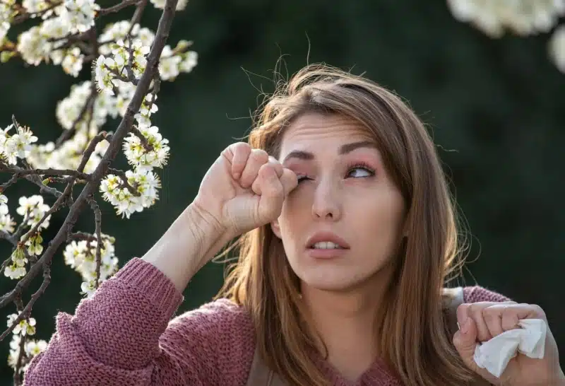 Young pretty girl with eye allergies rubbing eyes besides a blooming tree in spring time