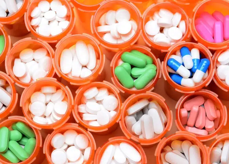 Looking down on a large group of prescription medicine bottles. The bottles all have their caps off and have a variety of drugs, tablets and capsules. Horizontal format filling the frame.