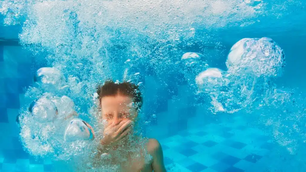 Little boy holding his breath covering his nose in a pool surrounded by bubbles