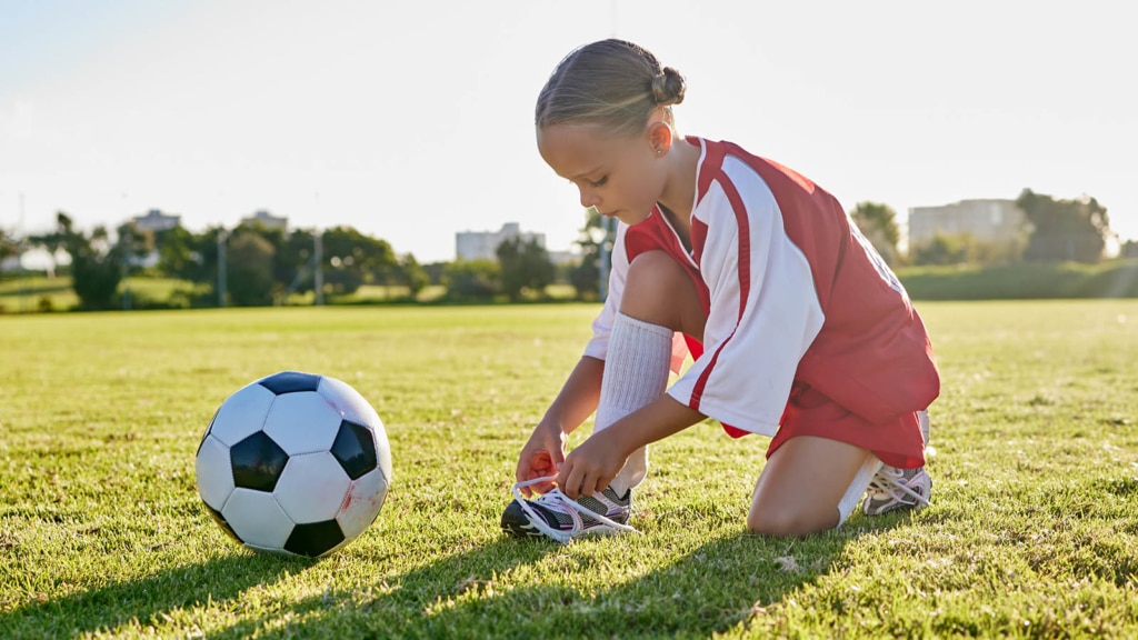 Little girl in a red soccer uniform tying her tennis shoe next to a soccer ball
