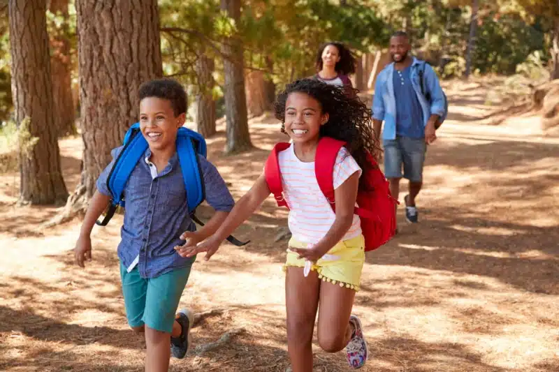 A brother and a sister running in the woods, both with backpacks and their parents walking behind them