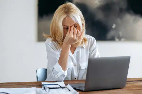 A mature blonde woman in an office with her laptop grabbing the start of the nose between her eyebrows with two fingers due to occupational asthma complications