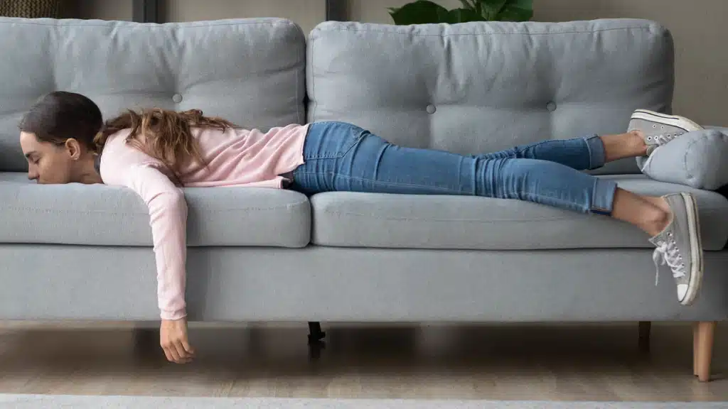 Young woman with pink shirt, jeans and tennis shoes, laying down on her belly on a gray couch showing signs of fatigue