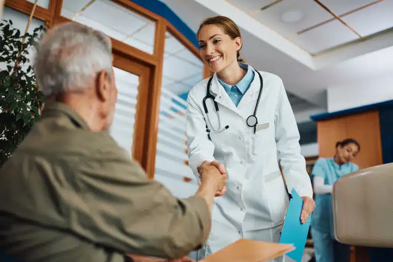 Happy female doctor and mature patient shaking hands while greeting in waiting room at medical clinic