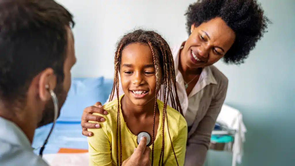 Male physician performing a health checkup with a stethoscope on a girl, her mother is besides her