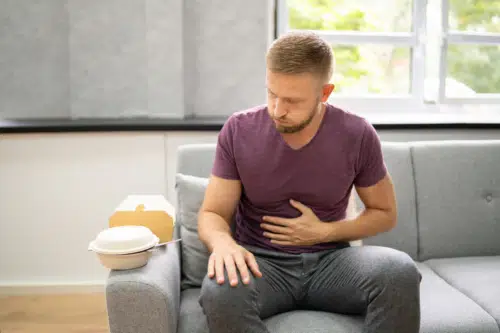 Man in a living room sitting on a couch holding his stomach in pain