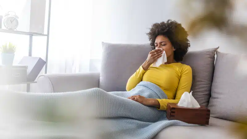 Sick young woman sitting on sofa blowing her nose at home in the sitting room.