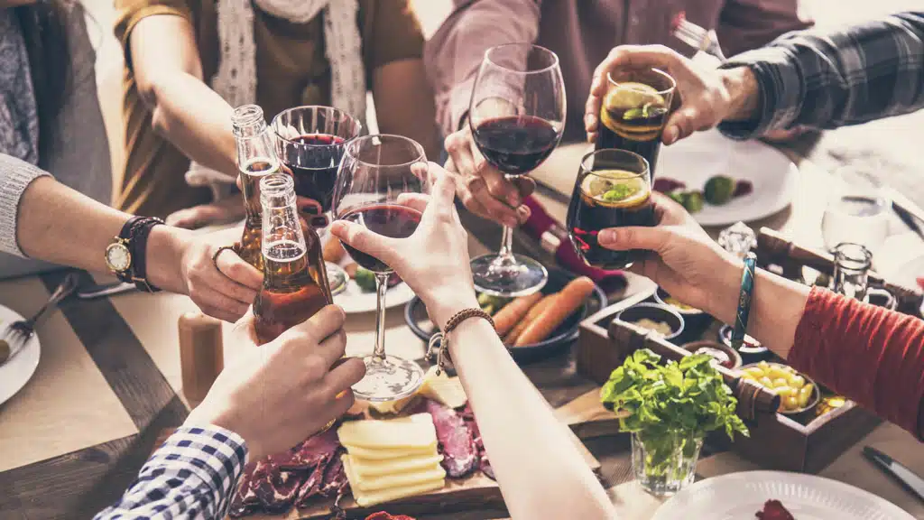 Group of friends cheering with different types wine, beer and cocktails over a table of cold cuts, veggies and other foods