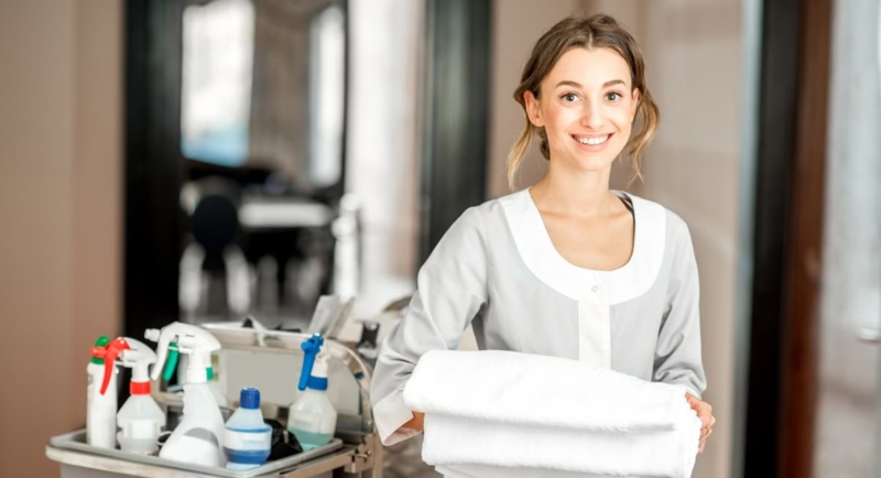 Housekeeper in a hotel corridor holding a towel standing with a cart full of cleaning products that can be irritants and cause occupational asthma