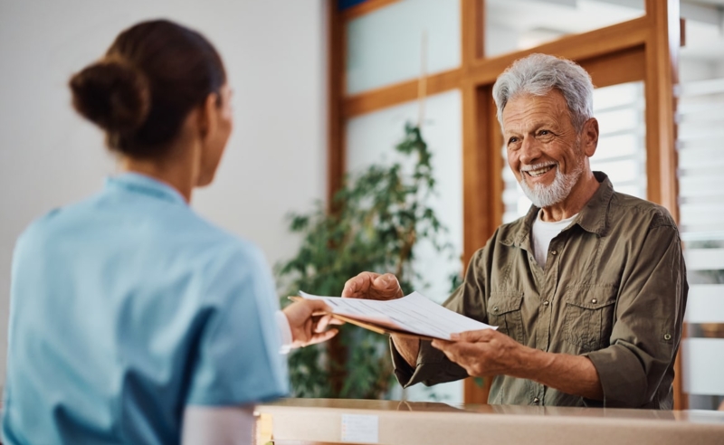 Happy senior man receiving his medical documents from nurse at reception desk at doctor's office.