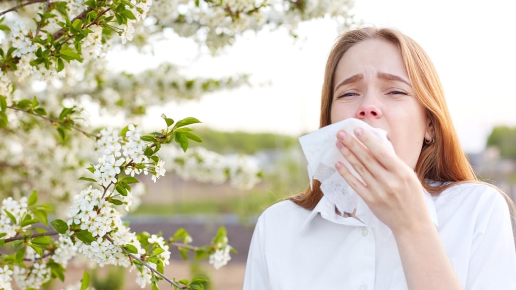 Woman outdoors next to flowers about to sneeze with a tissue paper in her hand 